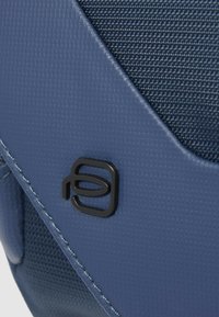 Blauwe stoffen tas met textuur, voorzien van een glad leren accent en een zwart logo detail, die een contrast in materialen en designelementen laat zien.