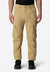 Beige vetoketjulliset housut, jotka on valmistettu kevyestä kankaasta, varustettuna taskuilla, sileällä pinnalla ja logolla reiden kohdalla. Yhdistetty mustiin kengiin.