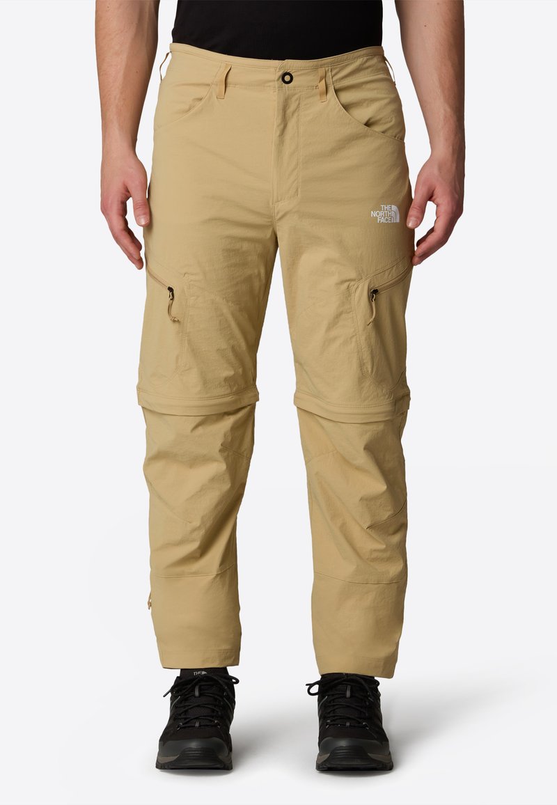 Beige vetoketjulliset housut, jotka on valmistettu kevyestä kankaasta, varustettuna taskuilla, sileällä pinnalla ja logolla reiden kohdalla. Yhdistetty mustiin kengiin.