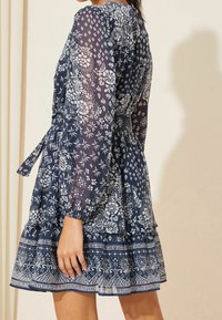 Woman wearing a navy blue floral patterned dress with long sheer sleeves and a tiered skirt, standing against a beige background.