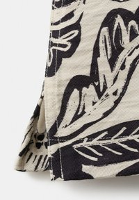 Gros plan d’un tissu beige avec un motif abstrait de feuilles et de branches noires, montrant l’ourlet cousu et le détail de la fente latérale.