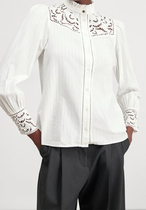 Blouse blanche à manches longues avec des détails en dentelle sur le haut de la poitrine et les poignets, boutonnée sur le devant, associée à un pantalon noir ample, mains dans les poches.