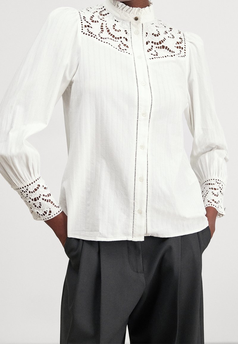 Blouse blanche à manches longues avec des détails en dentelle sur le haut de la poitrine et les poignets, boutonnée sur le devant, associée à un pantalon noir ample, mains dans les poches.