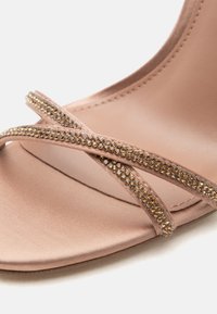 Κοντινό πλάνο γυμνού σατέν σανδαλιού με δύο λεπτά λουράκια διακοσμημένα με μικρές χρυσές στρας στην περιοχή των δακτύλων.