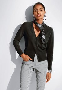 Schwarze Strickjacke mit V-Ausschnitt, Knopfverschluss und gerippten Bündchen, kombiniert mit gestreiften Hosen. Accessoires mit einem schwarz-weißen Schal.