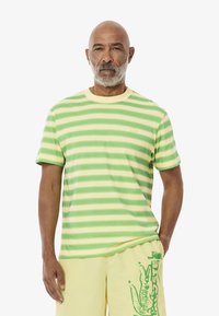 Gestreept T-shirt met groene en gele kleuren, ronde hals, korte mouwen en een klein logo op de linkerkant van de borst. Katoenen materiaal.