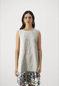 Mulher com cabelo escuro e longo, vestindo um vestido branco sem mangas com espirais texturizadas e uma barra de lantejoulas prateadas brilhantes, de pé contra um fundo simples.