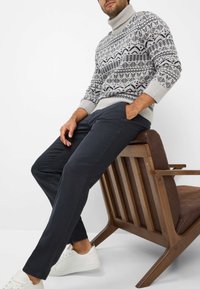 Gebreide trui met geometrische navy en grijze patronen; gecombineerd met donkere broeken en witte sneakers; zittend op een houten stoel.