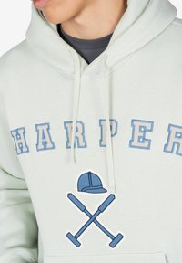 Sudadera verde claro con bordado en azul y beige que dice "HARPER". El gráfico central incluye un sombrero y palas cruzadas en azul. Textura suave.