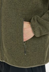 Olivgrön fleecejacka med texturerad yta, utrustad med en zip-försedd sidoficka och lättviktsmaterial. Accentuerad med matchande kant.