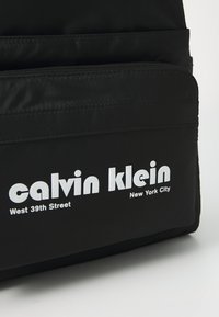 Sac en nylon noir avec deux compartiments zippés. Comprend du texte blanc : "calvin klein," "West 39th Street," et "New York City." Texture lisse.
