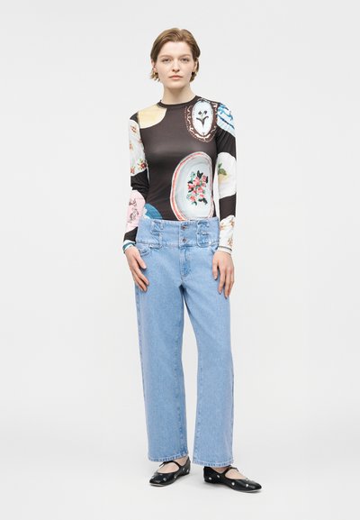 Γυναίκα που στέκεται, φορώντας μακρυμάνικο μαύρο τοπ με οβάλ λουλουδάτα σχέδια, τζιν ψηλόμεσο ανοιχτού γαλάζιου χρώματος και μαύρα επίπεδα παπούτσια με λουράκια.