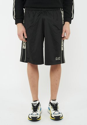 Persona che indossa pantaloncini neri EA7 con fasce laterali beige e sneaker EA7 brandizzate in bianco, nero, giallo e blu, in piedi su sfondo bianco.