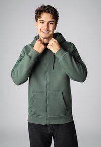 Grüne Zip-Hoodie aus weichem Material, mit Kapuze, frontalen Taschen und gerippten Bündchen. Sublevel-Logo auf der Vorderseite gestickt.