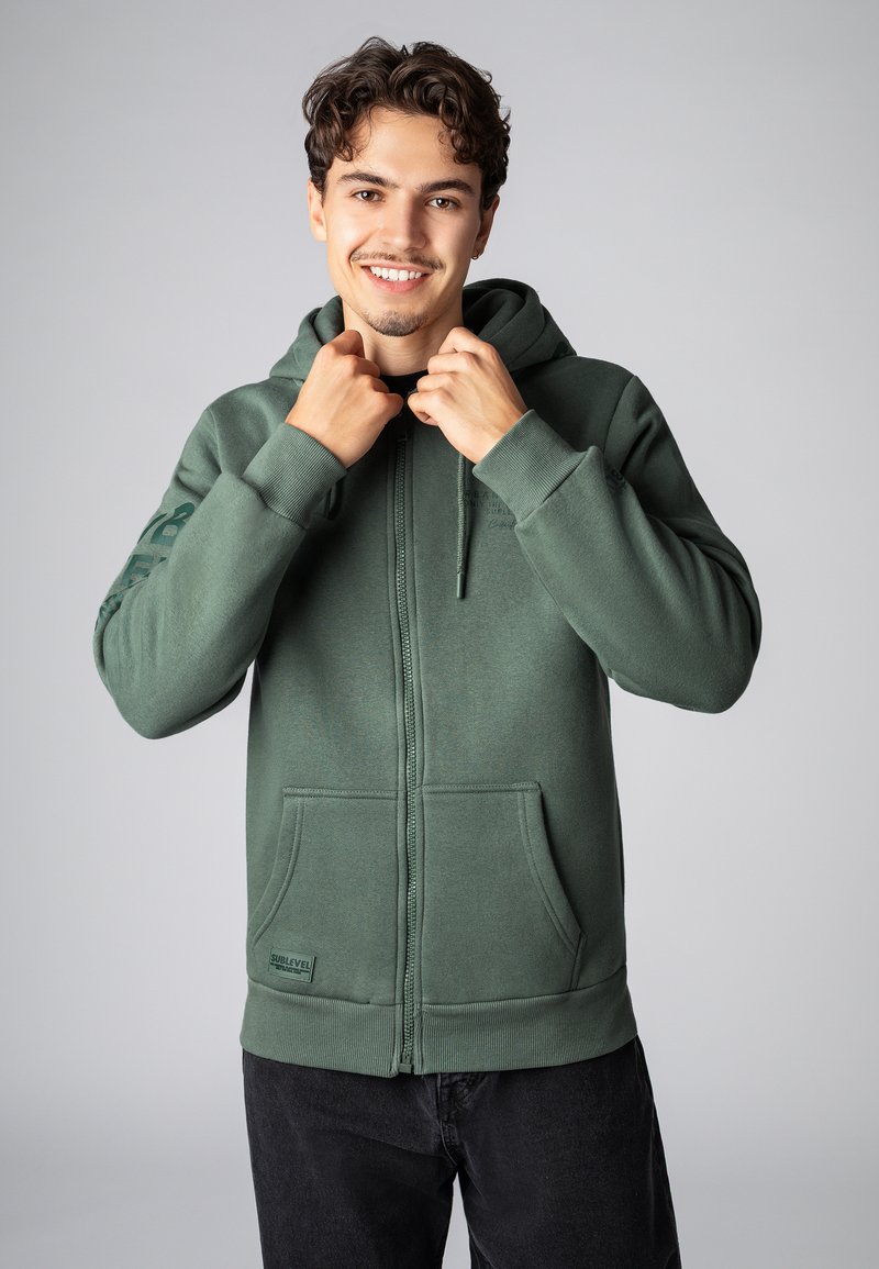Grüne Zip-Hoodie aus weichem Material, mit Kapuze, frontalen Taschen und gerippten Bündchen. Sublevel-Logo auf der Vorderseite gestickt.