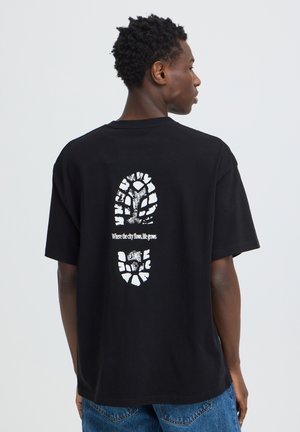 Homme portant un t-shirt noir avec une empreinte de botte blanche et le texte "Where the city flows, life grows" dans le dos, associé à un jean bleu.