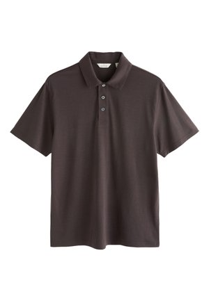 Uomo di mezza età con testa calva e barba grigia che indossa una polo marrone scuro, guardando frontalmente su uno sfondo semplice.