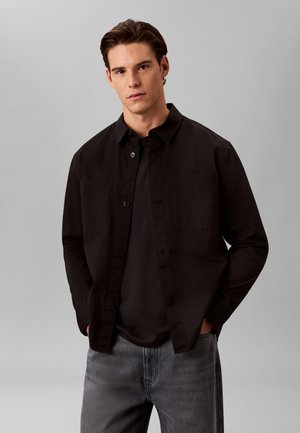 Νέος άνδρας με σκούρα μαλλιά, που φοράει ένα μαύρο πουκάμισο με κουμπιά πάνω από ένα μαύρο μπλουζάκι και γκρι τζην, στέκεται μπροστά σε ένα απλό ανοιχτό γκρι φόντο.