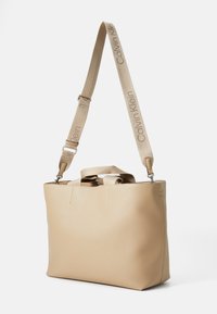 Beige skinnveske med doble håndtak, justerbar stropp og preget logo på stroppen. Glatt tekstur, strukturert design og minimal søm.