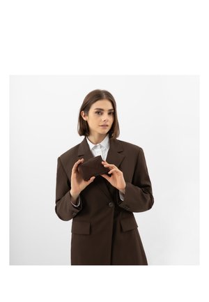 Vrouw in een bruine blazer die met beide handen een bijpassende bruine portemonnee vasthoudt tegen een effen witte achtergrond.