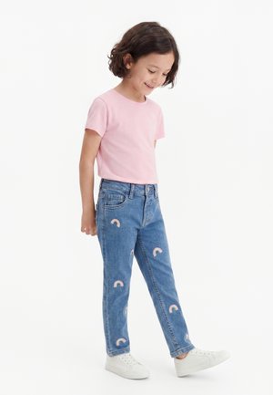 Jeune fille en t-shirt rose et jean bleu avec de petits patchs arc-en-ciel, souriante et regardant vers le bas, portant des baskets blanches, debout sur fond blanc.