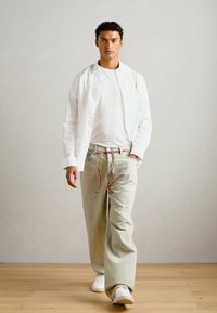 White shirt over a white t-shirt, light green trousers with drawstring, and white sneakers, standing on a wooden floor against a neutral background.