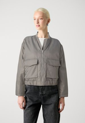 Grijze cropped bomberjack met lange mouwen, ritssluiting aan de voorkant, twee voorkennis en een elastische zoom. G gedragen met donker jeans en een witte top.