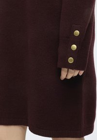 Gros plan d'une personne portant un vêtement en maille bordeaux foncé avec trois boutons décoratifs dorés sur le poignet de la manche.