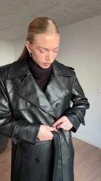 Cappotto doppiopetto in pelle nera con grandi risvolti, dettagli con bottoni e cintura. Indossato sopra un maglione lavorato a maglia scuro, che mette in risalto il tessuto strutturato.