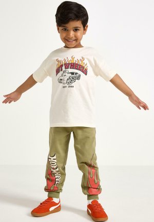 T-shirt crema con grafiche di fiamme rosse e gialle e illustrazione di un'auto, abbinato a jogger oliva con design di fiamme simili e sneakers arancioni.