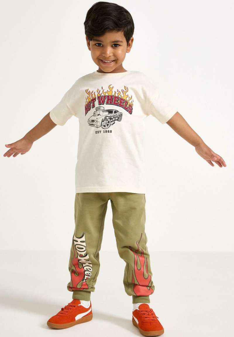 Crème T-shirt met rode en gele vlamgraphics en auto-illustratie, gecombineerd met olijfgroene joggers met vergelijkbare vlamontwerpen, en oranje sneakers.