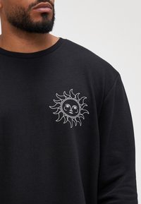 Camisa negra de manga larga con un diseño de sol bordado en blanco en el lado izquierdo del pecho. Tejido suave con un corte relajado y cuello redondo.