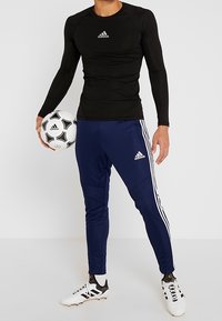 Black long-sleeve athletic shirt and navy track pants with white stripes. Holding a black and white soccer ball, wearing white cleats.