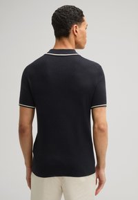 Polo navy con tessuto testurizzato, maniche corte, colletto bianco e dettagli bianchi sulle maniche. Mostrato da dietro, evidenziando la vestibilità e i dettagli di design.
