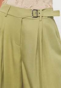 Ανοιχτό πράσινα ψηλόμεσα παντελόνια με φαρδύ πόδι, πιέτες και αντίστοιχο ζωνάκι με ορθογώνια ασημένια αγκράφα και ραφές.
