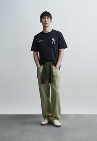 Μαύρο κοντομάνικο μπλουζάκι με μοβ καρτούν γραφικό, σε συνδυασμό με ανοιχτό πράσινο ριγέ παντελόνι και λευκά αθλητικά παπούτσια. Λεπτό πράσινο πουλόβερ δεμένο στη μέση.