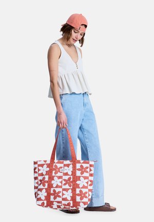 Joven con gorra coral, top blanco sin mangas y vaqueros azul claro, sosteniendo un gran bolso tote con estampado floral rojo y blanco.