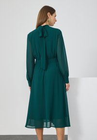 Robe longue semi-transparente teal à manches longues avec un nœud dans le dos et une taille élastique. Elle présente des poignets boutonnés et une jupe évasée fluide.