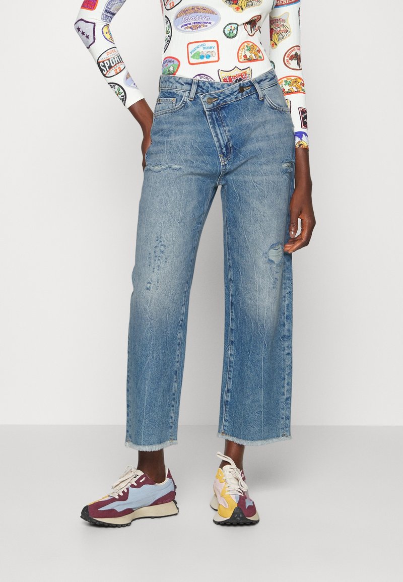 Personne portant un jean taille haute à jambes larges déchiré avec des ourlets effilochés, un top vintage coloré à manches longues avec patchs, et des baskets multicolores.