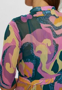 Rochie transparentă cu un model abstract multicolor vibrant, incluzând nuanțe de roz, verde, galben și albastru. Are un guler și un detaliu cu talie accesorizat cu brâu.