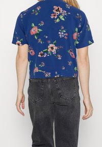 Person iført en blå kortærmet skjorte med pink blomstermønster og højtaljede, falmede sorte jeans, set fra ryggen.