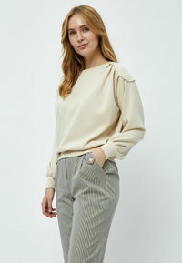 Felpa beige a maniche lunghe con dettaglio sulle spalle arricciato, abbinata a pantaloni grigi con righe verticali, dotati di tasche frontali e bottone decorativo.