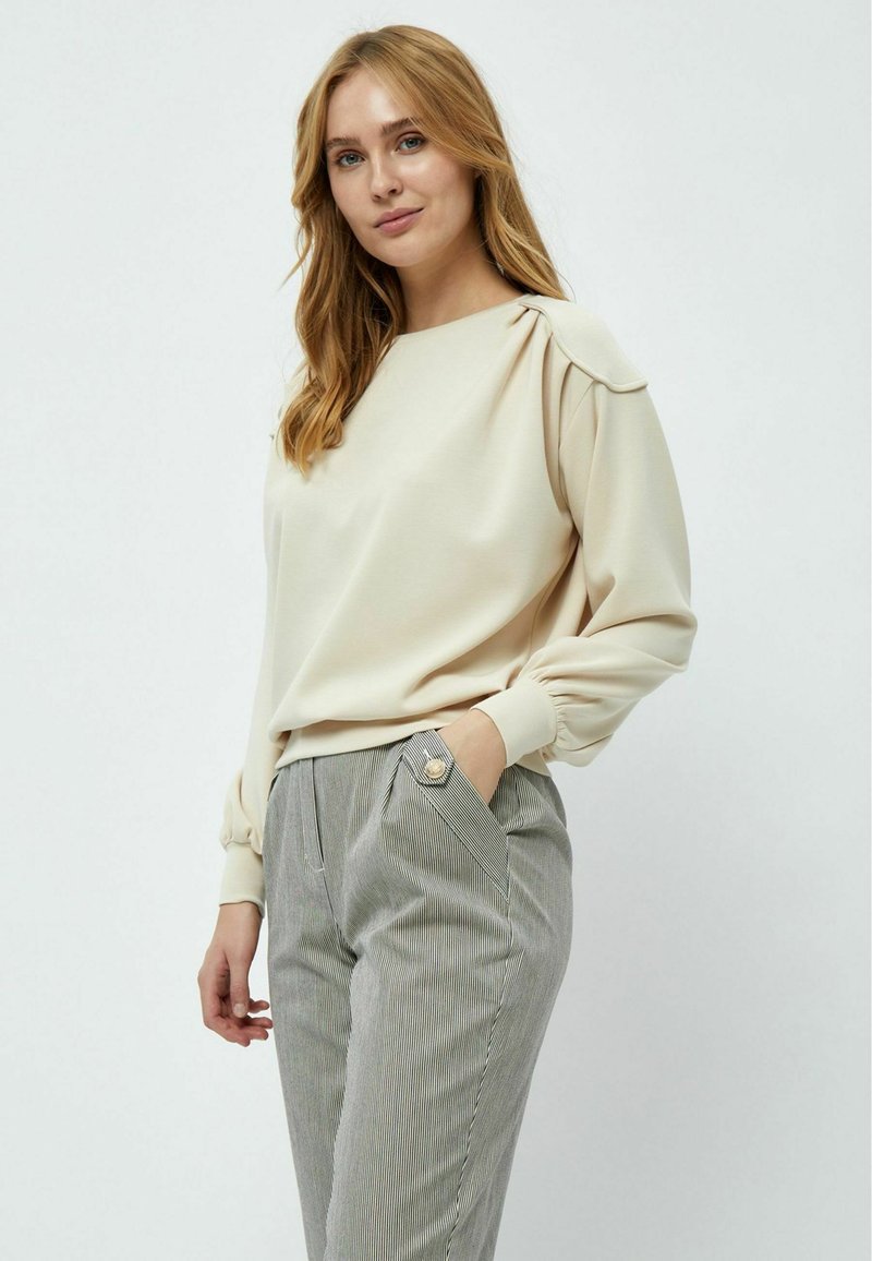 Felpa beige a maniche lunghe con dettaglio sulle spalle arricciato, abbinata a pantaloni grigi con righe verticali, dotati di tasche frontali e bottone decorativo.