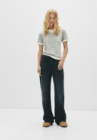 Grå t-shirt med rund hals och vit kant, i kombination med mörka denimbyxor med utsvängda ben. Modellen bär beiga slip-on skor och silverringar.