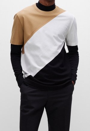 Farbblocking-T-Shirt mit Abschnitten in Beige, Weiß und Schwarz. Gerippter Kragen und strukturierter Stoff mit kurzen Ärmeln, über einem schwarzen Shirt getragen.