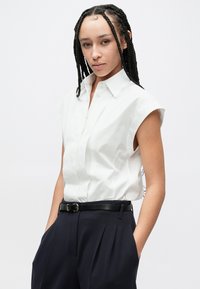 Άσπρο πουκάμισο με κοντό μανίκι και κουμπιά, με δομημένο γιακά, συνδυασμένο με μπλε σκούρο παντελόνι ψηλής μέσης και μαύρη ζώνη. Μινιμαλιστικός σχεδιασμός.