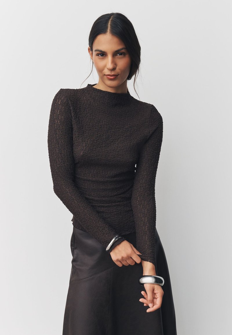 Haut à manches longues texturé marron avec un col montant, associé à une jupe en cuir noire. Bracelets argentés aux poignets.