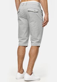 Homme portant un short gris à carreaux jusqu'aux genoux et des baskets blanches, vu de dos sur un fond uni.