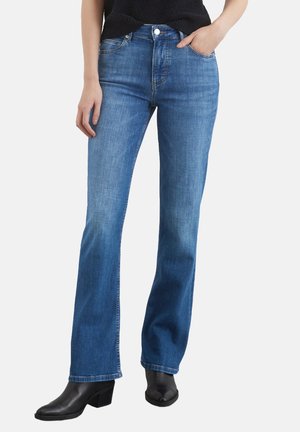 Femme portant un jean bleu taille moyenne légèrement évasé et des bottines noires, main dans la poche, sur fond blanc uni.