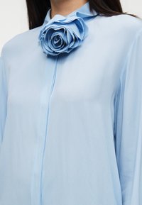 Ανοιχτό μπλε μπλούζα από διαφανές ύφασμα, με μεγάλη υφασμάτινη διακοσμητική τριανταφυλλιά στον γιακά. Ομαλή υφή με κουμπιά στο εμπρός μέρος.